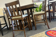 Juego de comedor alto en madera oscura con mesa cuadrada y cuatro sillas con respaldo vertical y asientos beige. El conjunto está exhibido en una tienda de muebles junto a plantas decorativas y otros comedores.