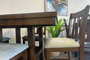 Detalle de juego de comedor alto en acabado madera oscura con sillas de respaldo vertical y asientos acolchados beige. El conjunto está exhibido en una tienda de muebles con planta decorativa al fondo.