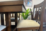 Primer plano de mesa alta de comedor en madera oscura con silla de respaldo vertical y asiento acolchado color beige. El conjunto está exhibido en una tienda de muebles con planta decorativa y arte mural al fondo.