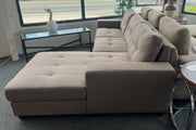 Brown tufted fabric sectional sofa with left-facing chaise lounge on dark carpet in showroom.