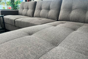 Gray tufted fabric sectional sofa with chaise lounge, woven texture, button details, near window with plant.