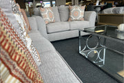 Sala de exhibición con sofá y loveseat tapizados en tela gris claro, cojines decorativos con patrones geométricos en tonos beige y terracota, y mesa de centro rectangular con estructura metálica y cubierta de vidrio.