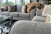 Sala de exhibición con sofá y loveseat tapizados en tela gris claro, cojines decorativos con patrones circulares en tonos beige y terracota, y mesa de centro de vidrio con estructura metálica frente a grandes ventanas iluminadas.