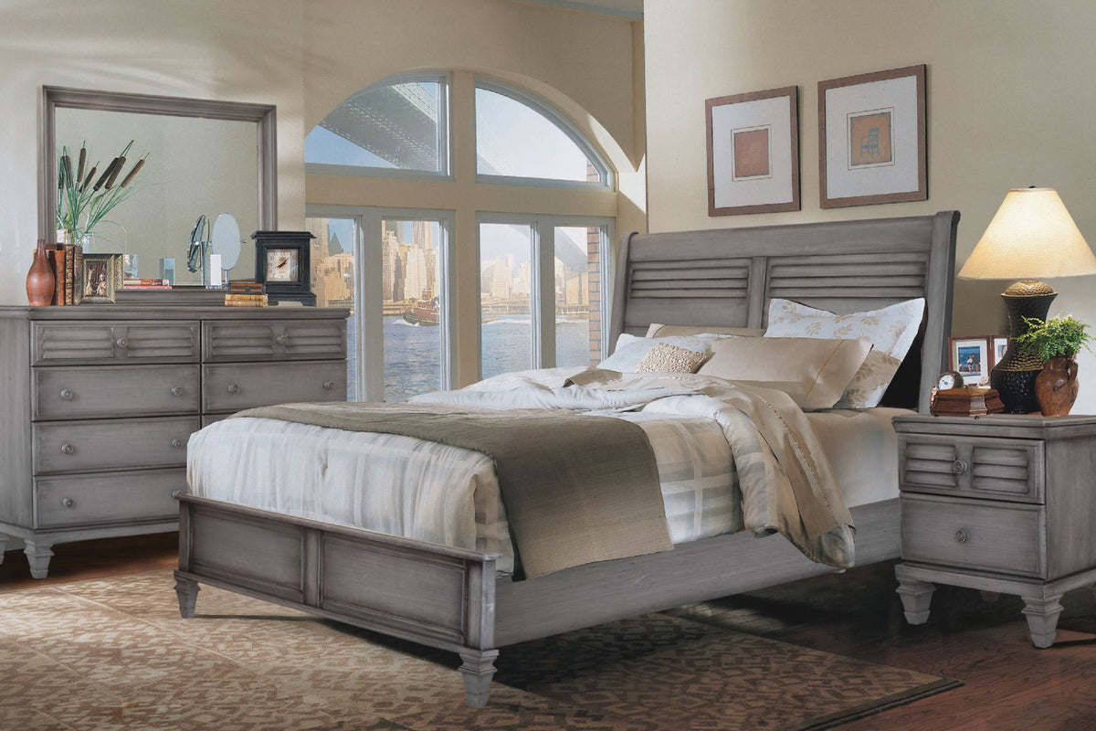 Conjunto de dormitorio de madera gris que incluye una cama, un gavetero con espejo y una mesa de noche en una habitación con una ventana que ofrece vistas a la ciudad.