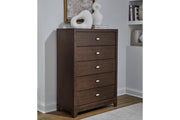 Chest marrón oscuro de cinco gavetas con tiradores ovalados plateados y elementos decorativos en su superficie, en una decoración de habitación.